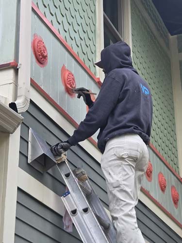 Man with dark grey hoodie on a ladder painting the exterior of a home.