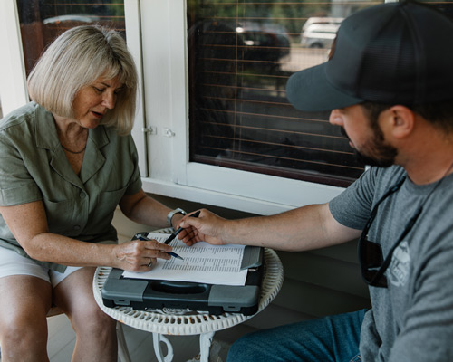Woman in her 60s with a bob haircut discussing home repairs with white man in his 30s. They have paperwork on the table in front of them.