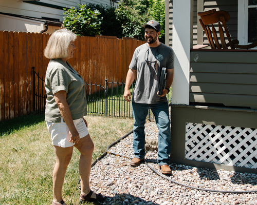 Woman in her 60's with a bob haircut talking to a man in his 30s with a dark beard. They are standing outside a home discussing home repairs.